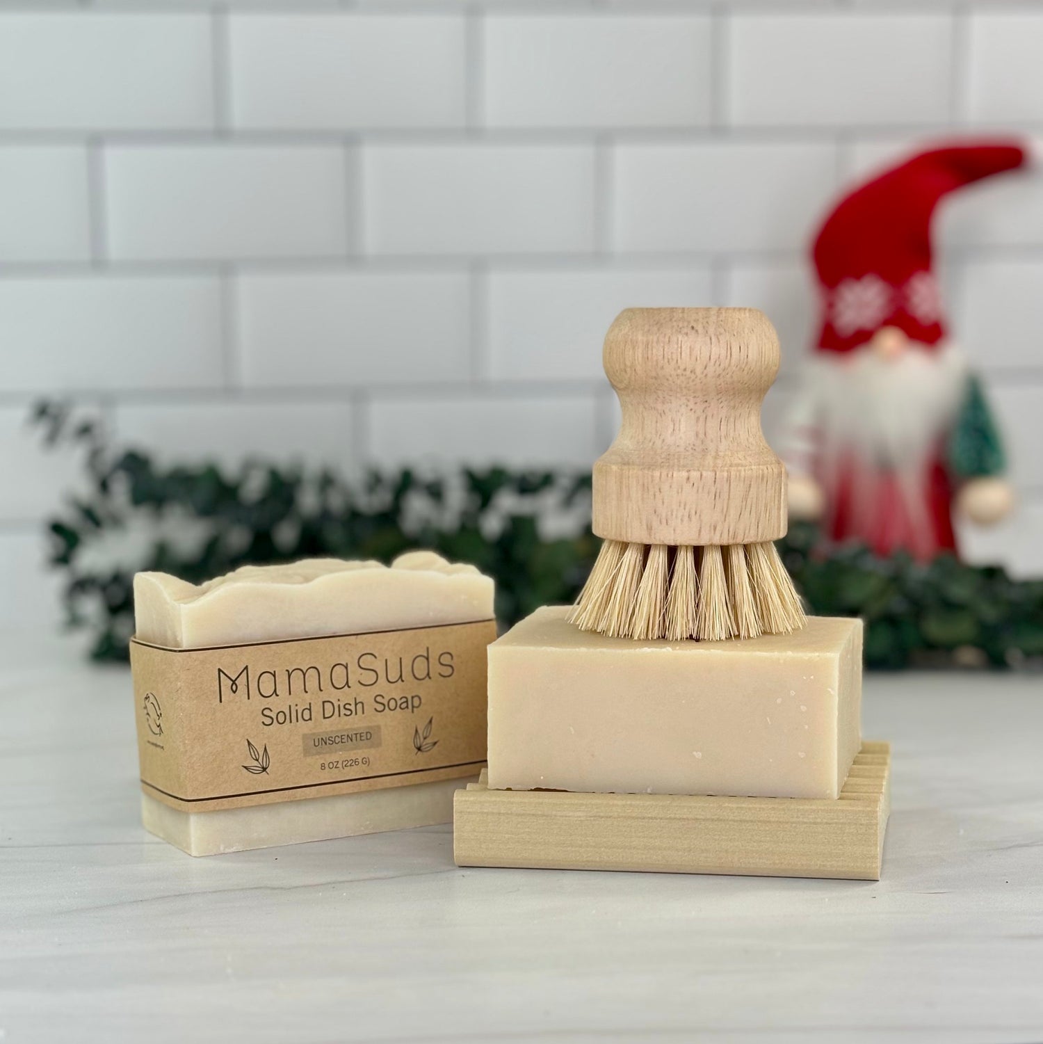 Solid Dish Soap in paper label standing next to a pot brush stacked on top of a solid dish soap on top of a wooden soap dish on a kitchen counter top in front of a eucalyptus sprig and a holiday gnome.